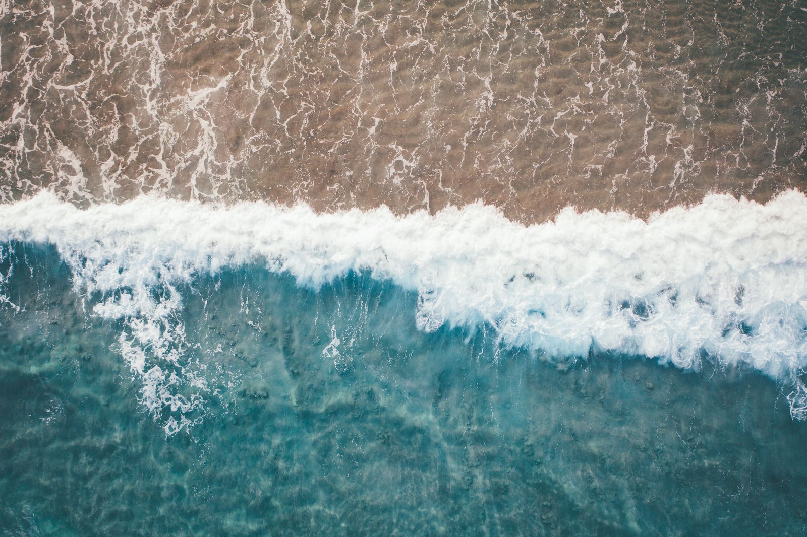 A wave breaking onto brown sand, its white line seen from directly above